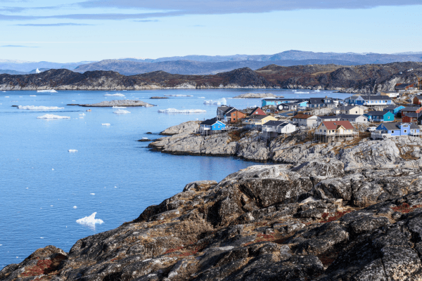 Coastal view of Ilulissat, Greenland, with colourful houses along rocky shoreline beside Disko Bay, dotted with drifting icebergs.